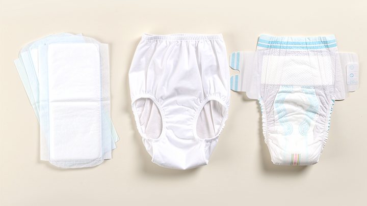 Symbolbild: Drei Formen der aufsaugenden Versorgung nebeneinander: ein Stapel Inkontinenzvorlagen, eine weiße Netzhose und eine Windelhose mit Klebestreifen